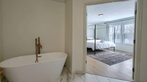 A modern bathroom with a freestanding bathtub, a glass shower enclosure, and a view of a bedroom with a bed and a window.