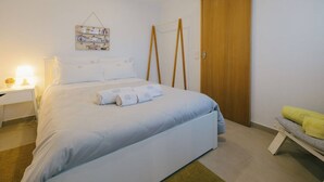 Een slaapkamer met een bed, een nachtkastje met een lamp, een houten garderobe en een bankje met een handdoek.