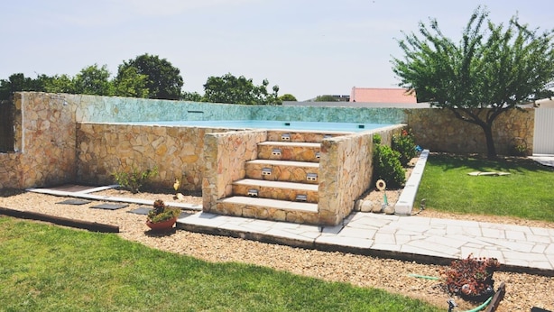A swimming pool with stone steps leading into it, surrounded by a stone wall and a grassy area with a tree.