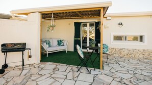 An outdoor patio area with a stone floor, a barbecue grill, a white sofa with cushions, a small table, and a hanging plant.