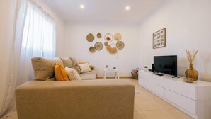 A modern living room with a beige sofa, a small side table, a TV on a white cabinet, and decorative wall art. - Tree House Casa Laranjeira (Sesimbra)