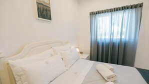 A neatly made bed with white linens and pillows, a small bedside table with a lamp, and a window with grey curtains. - Tree House Casa Laranjeira (Sesimbra)