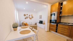 A modern kitchen with wooden cabinets, a gas stove, and a built-in oven. There is a dining area with a white table and chairs, and a small living space with a sofa and a TV. - Tree House Casa Laranjeira (Sesimbra)