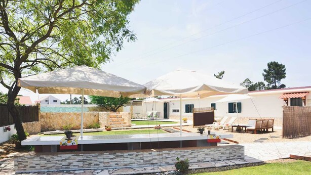 A backyard with a stone patio, outdoor furniture, and umbrellas providing shade. - Tree House Casa Laranjeira (Sesimbra)
