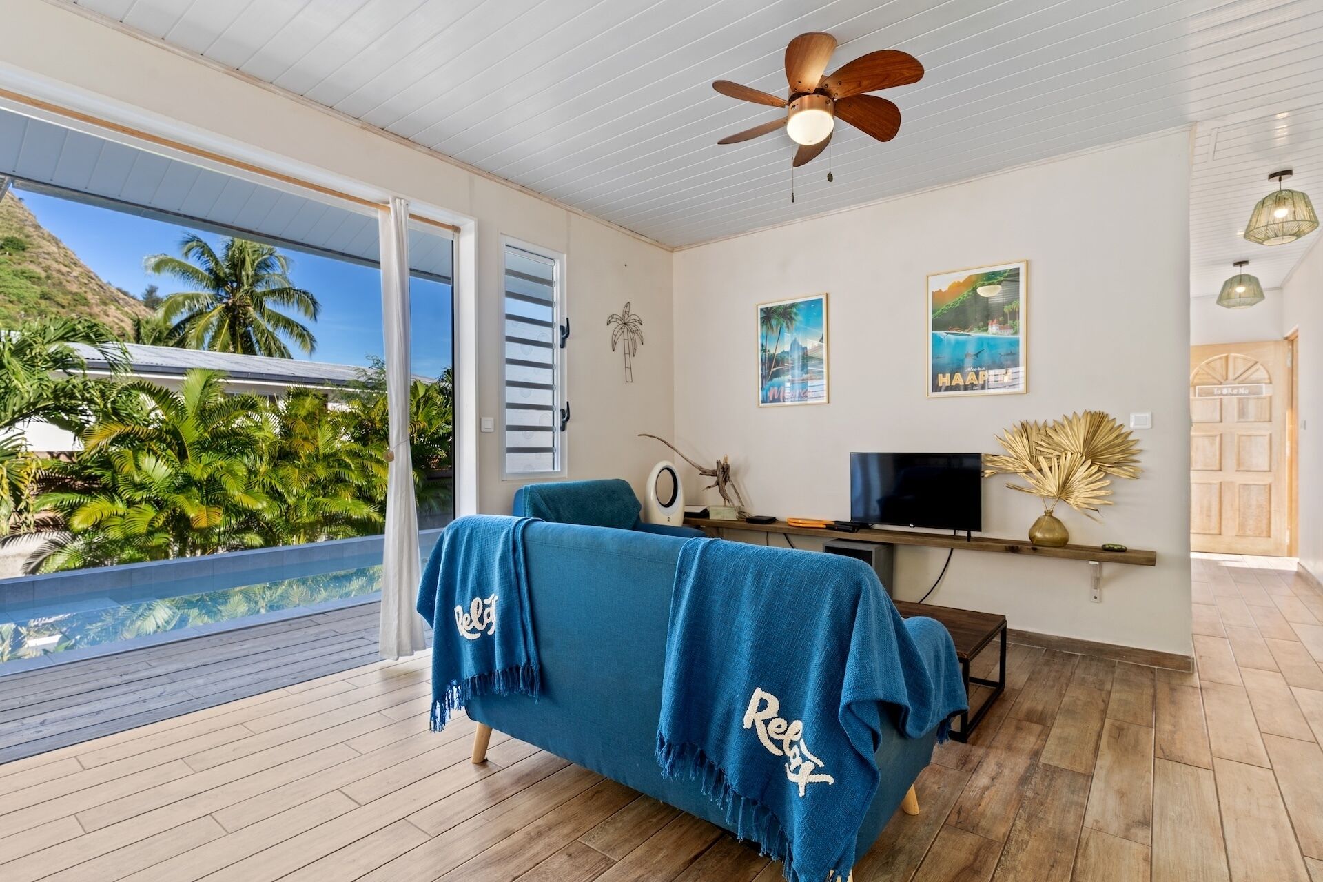Une pièce avec un lit recouvert d’une couverture bleue, un ventilateur de plafond, un téléviseur et un plancher en bois.