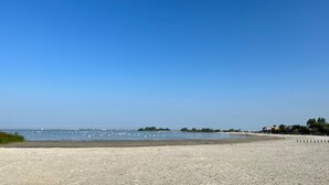 En strand med klar himmel, stilla vatten och en avlägsen kustlinje med byggnader.