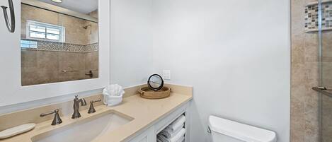 Un baño moderno con un espejo grande, una encimera color beige, un lavabo, un inodoro y una zona de ducha.
