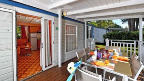 Un área de comedor al aire libre, techada, con una mesa preparada para dos personas, una pequeña cocina visible a través de una puerta abierta y vistas a un jardín.