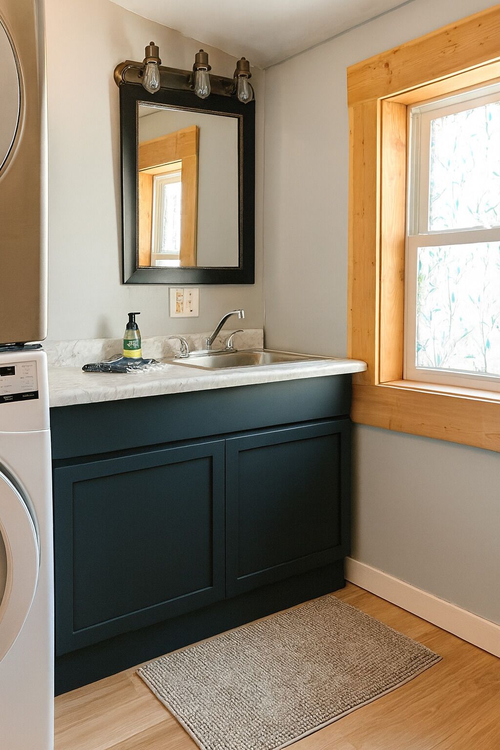 A bathroom with a dark blue vanity, a white sink, a mirror with three light fixtures, and a window with a wooden frame.