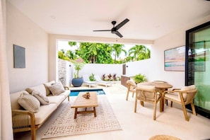 A modern living room with a sofa, coffee table, and wicker chairs. A ceiling fan is visible, and there is a view of greenery outside. - Azzurro #9 - by Zenbreak (Holetown)