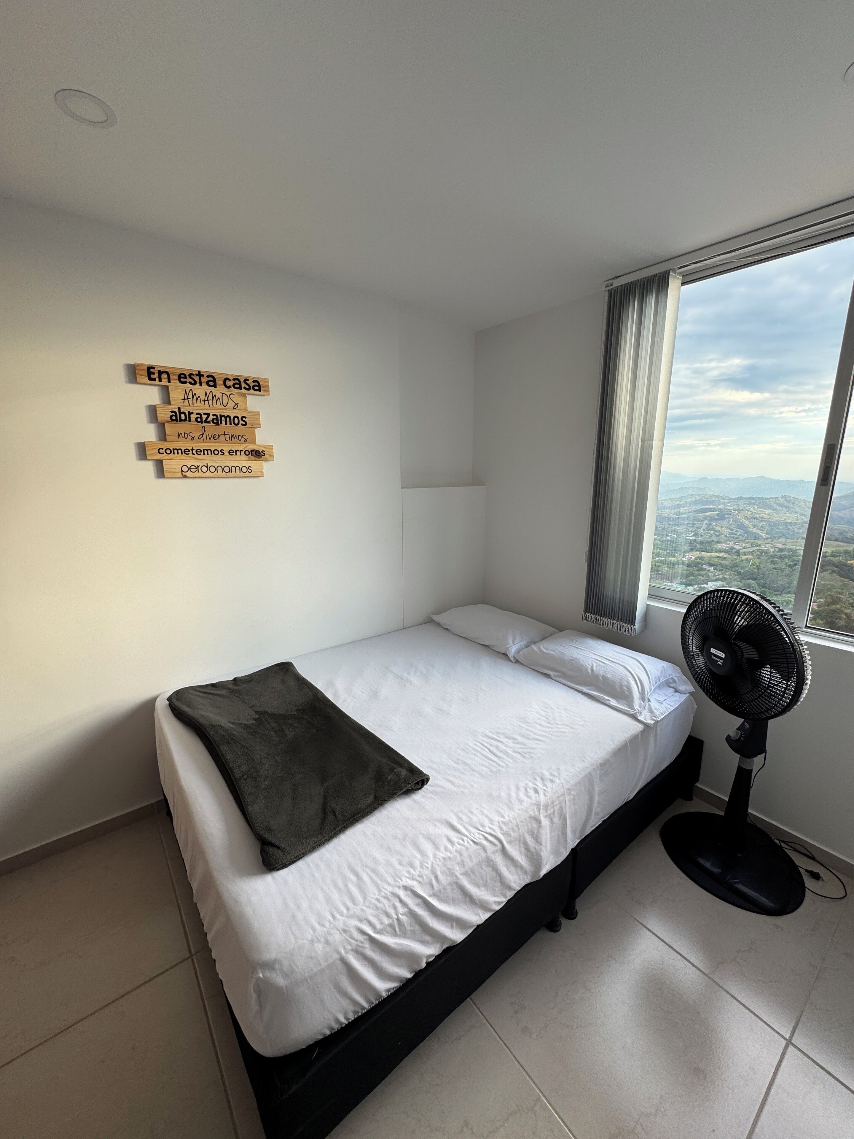 Une chambre avec un lit simple, une grande fenêtre, un ventilateur sur pied et un panneau fixé au mur avec du texte.
