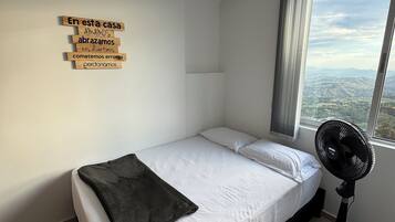 Une chambre avec un lit simple, une grande fenêtre, un ventilateur sur pied et un panneau fixé au mur avec du texte.