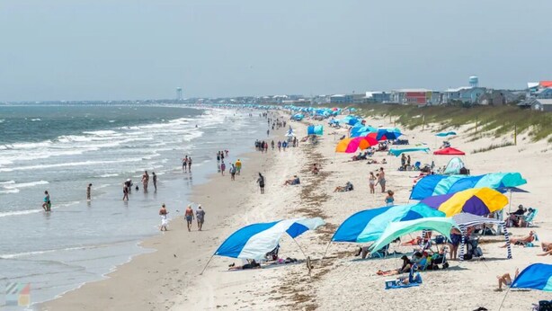 Ein Strand mit Zelten und Sonnenschirmen, Menschen die spazieren gehen, und eine Küstenlinie mit Gebäuden im Hintergrund.