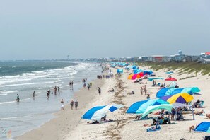 A beach with tents and umbrellas, people walking, and a coastline with buildings in the background.