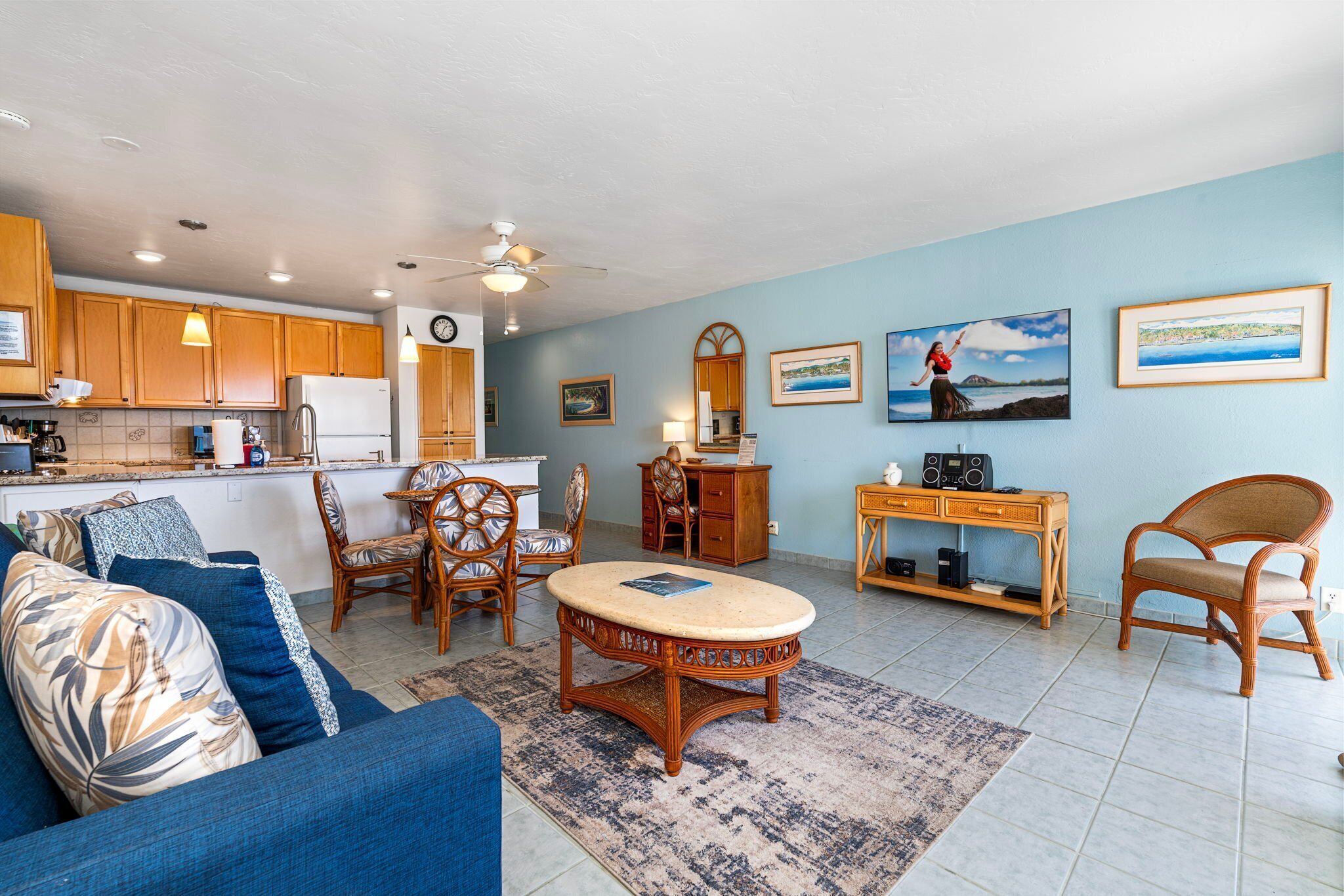 A spacious living area with a blue sofa, a round wooden coffee table, and a dining set. There is a kitchen in the background with wooden cabinets and a refrigerator.