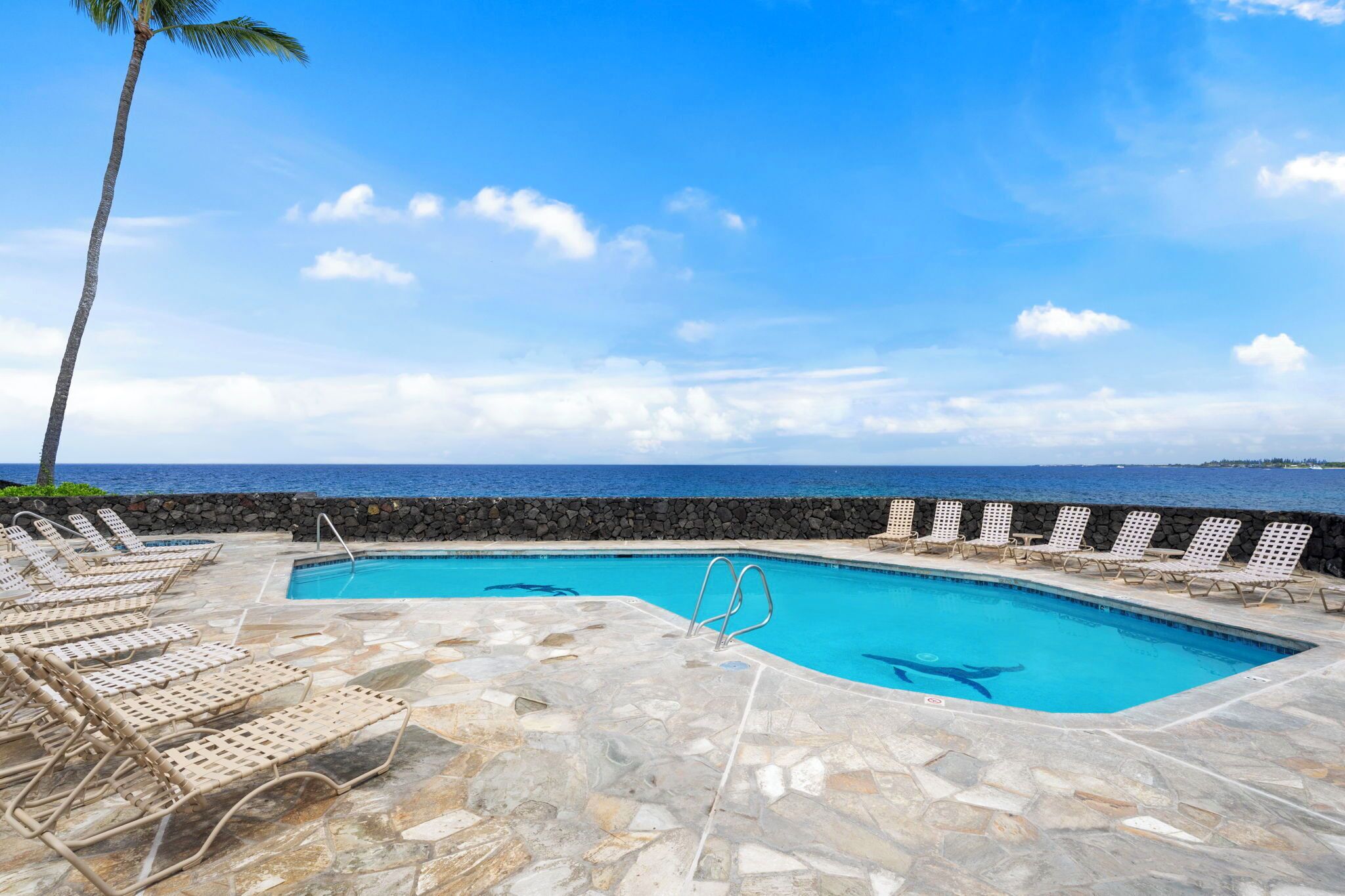 A pool area with lounge chairs and a stone wall, overlooking the ocean.