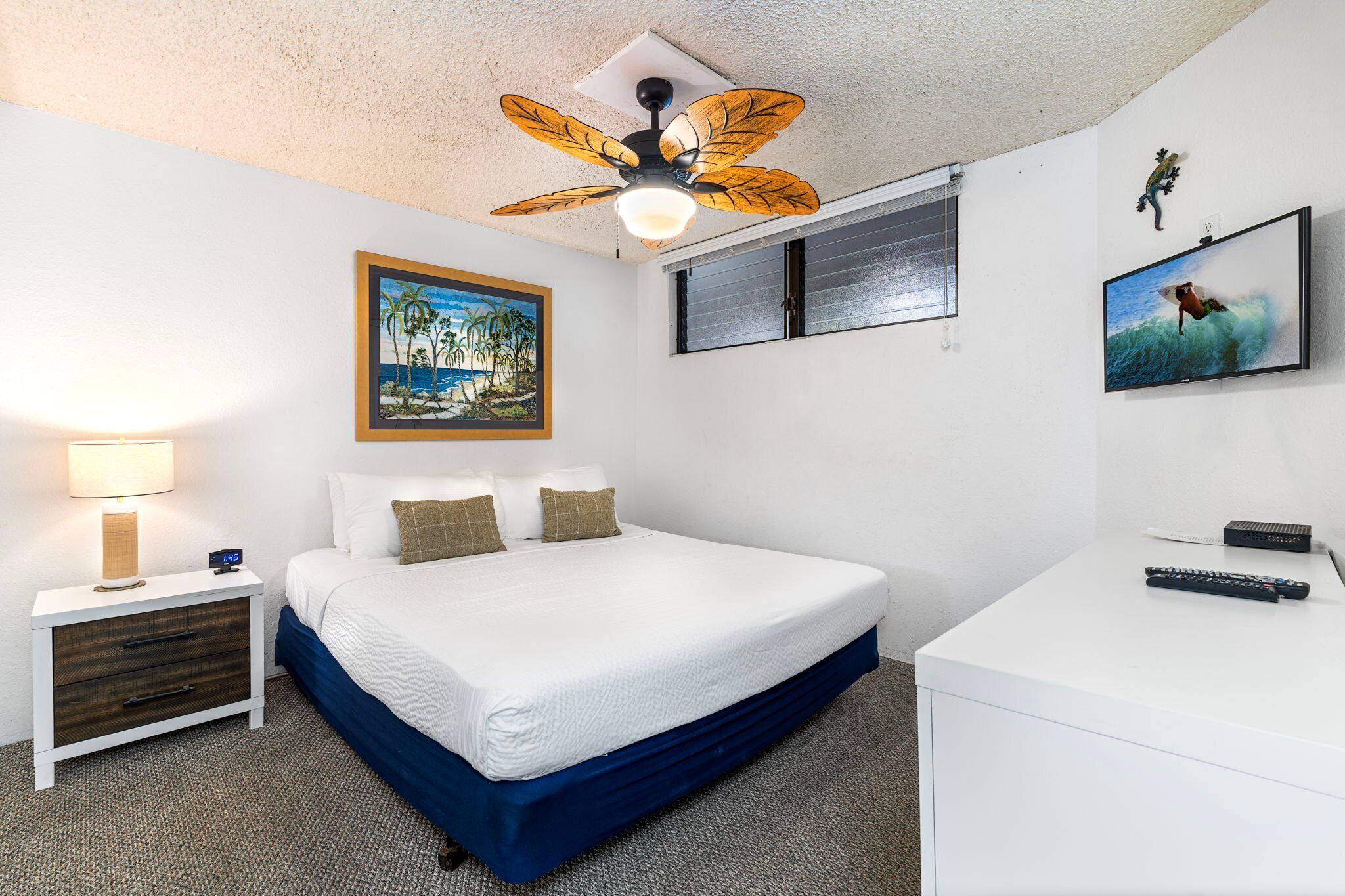A hotel room with a bed, a nightstand with a lamp, a ceiling fan, a TV, and a framed picture on the wall.