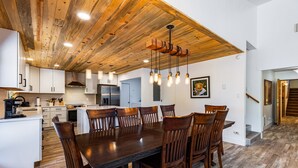 A spacious dining area with a large wooden table and chairs, a kitchen in the background, and a wooden ceiling.