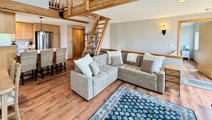 A spacious living area with a large sectional sofa, a dining table with chairs, a kitchen with modern appliances, and a wooden staircase. - Beachfront ADA Cottage: Elevator, Sauna, fire pit (Westport)