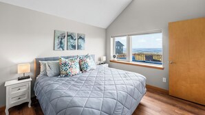 A bedroom with a large bed, a nightstand, a window with a view of the sea, and artwork on the wall. - Beachfront ADA Cottage: Elevator, Sauna, fire pit (Westport)