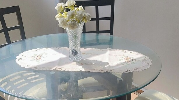 Una mesa redonda de cristal con mantel de encaje, rodeada de sillas de madera.