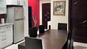 Un coin repas avec une table en bois foncé et des chaises noires, une cuisine moderne dotée d’un réfrigérateur en acier inoxydable et un mur d’accent rouge.