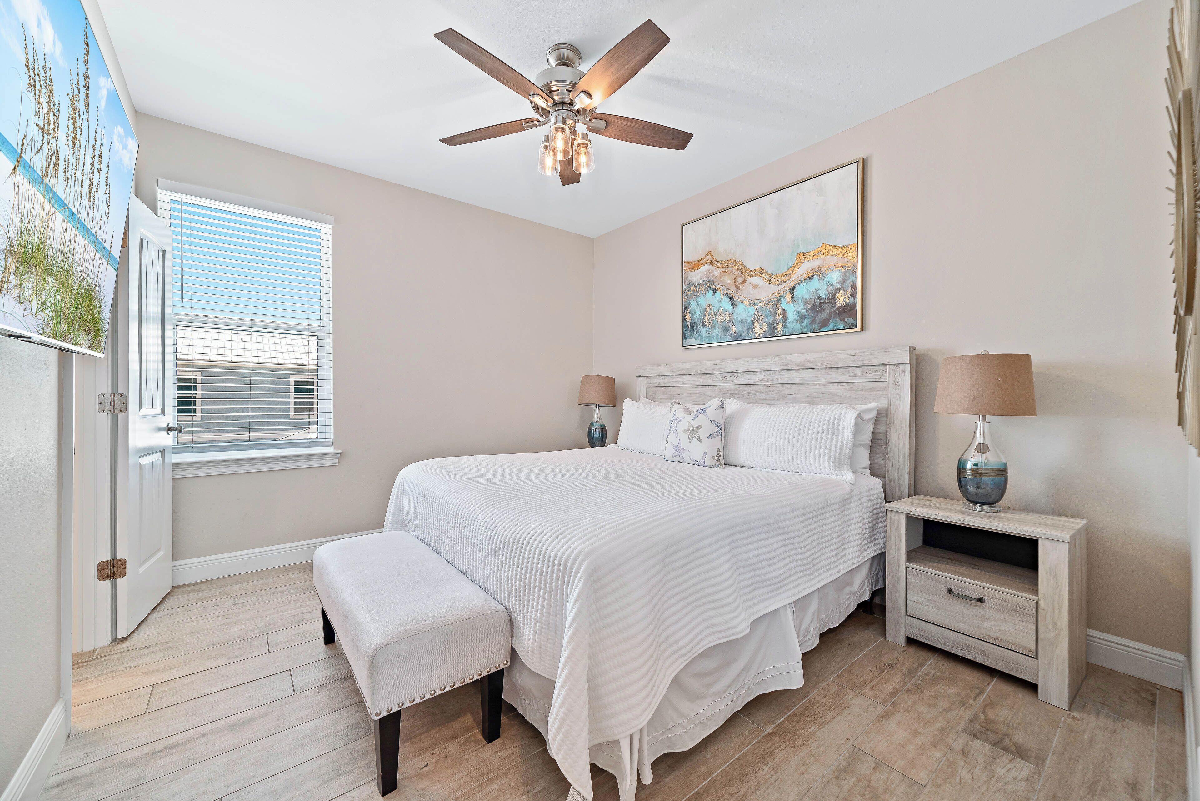 A bedroom with a bed, bench, nightstand, lamp, ceiling fan, window with blinds, and artwork on the wall.