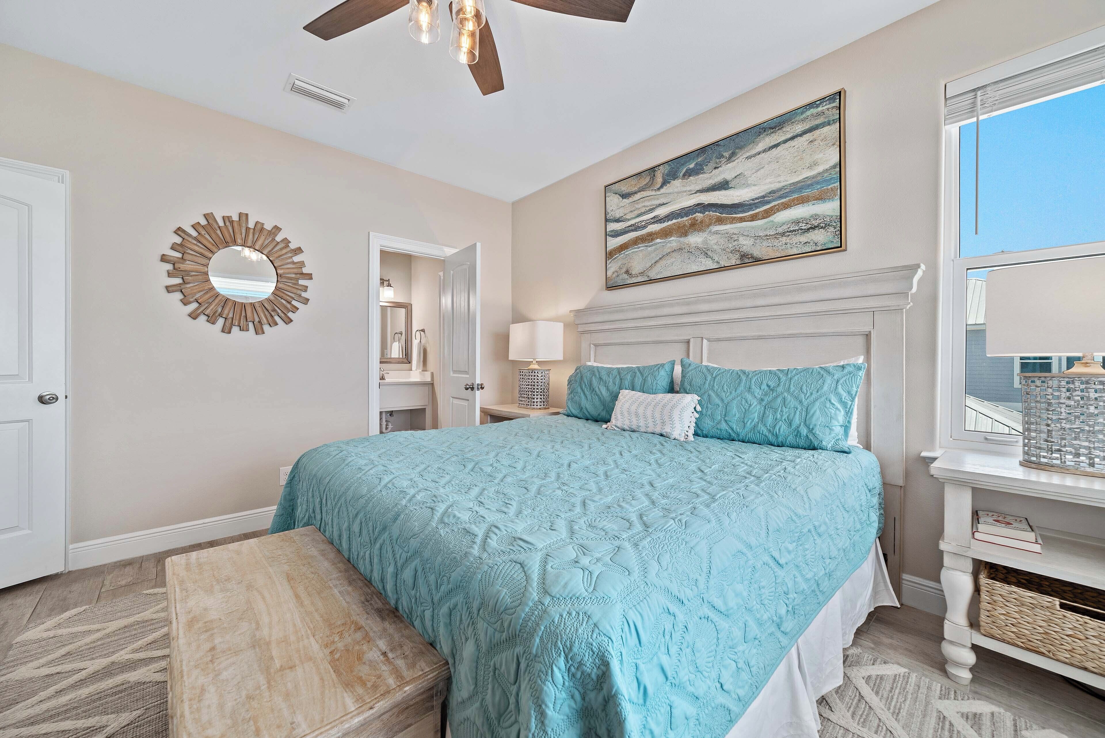 A bedroom with a bed, a ceiling fan, a wall-mounted mirror, a painting, and a window.