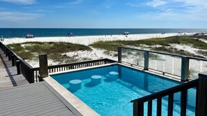 A pool with a wooden deck, a beach with sand dunes, and the ocean in the background.