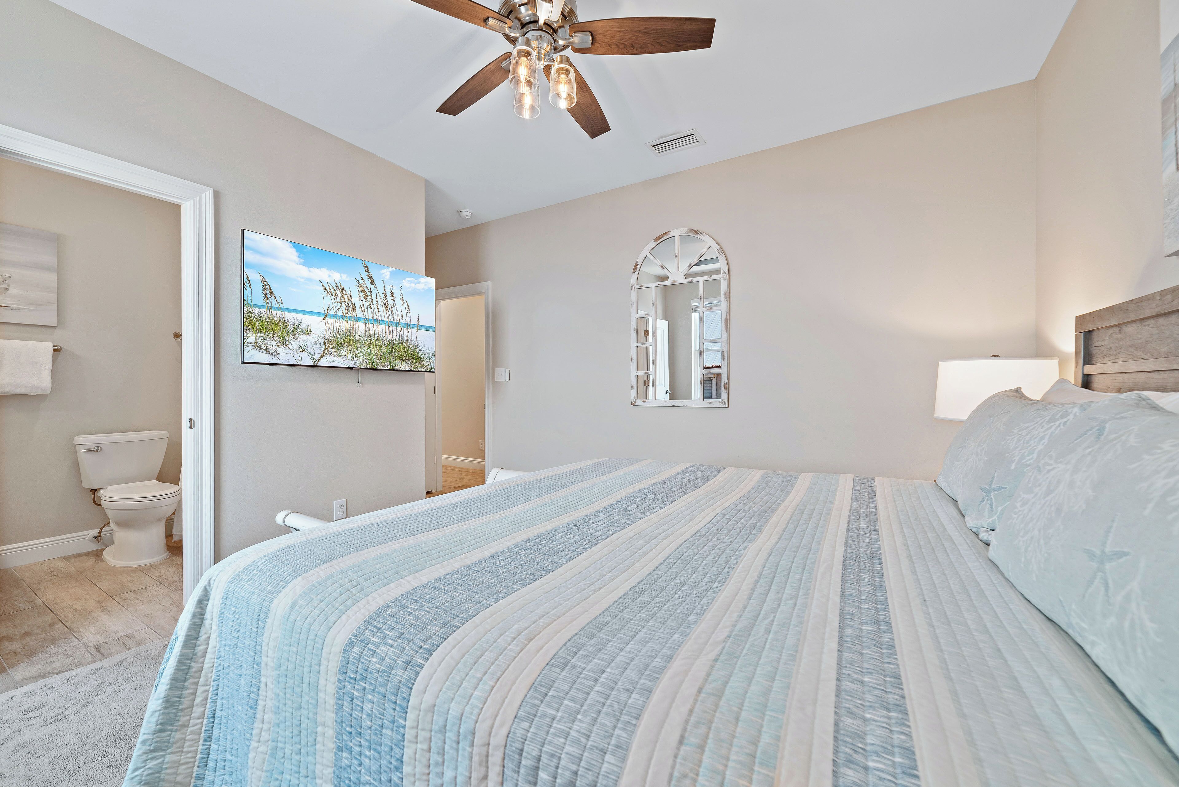A bedroom with a bed, a ceiling fan, a painting, a mirror, and a toilet.