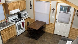 A compact kitchen with wooden cabinets, a stove, and a microwave. There is a dining table with chairs, a gray sofa, and a small wooden table. - Cozy Cabin w/ loft in charming Colorado Springs near USAFA (Colorado Springs)