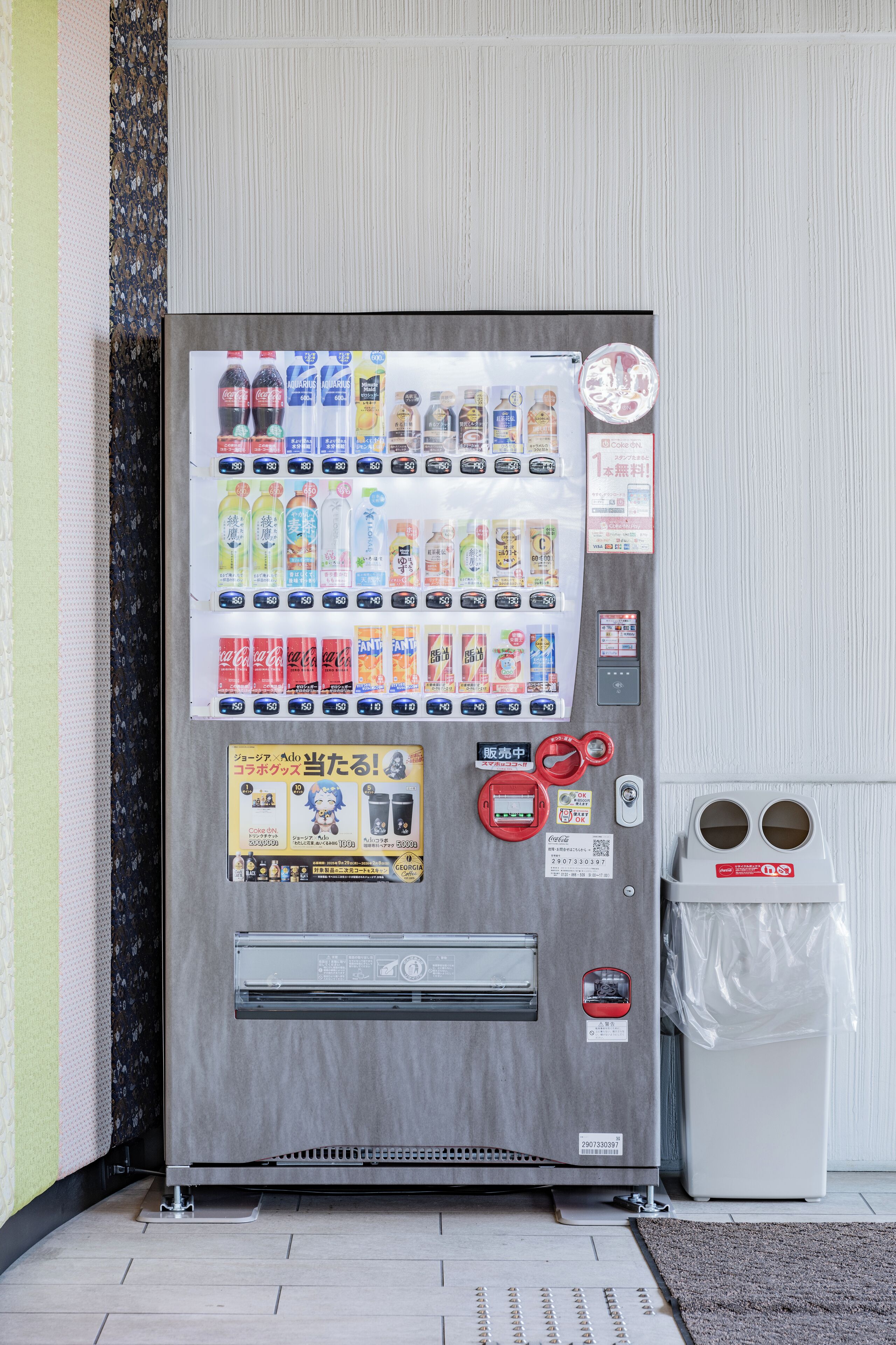 A vending machine with various drink options and a trash bin beside it.