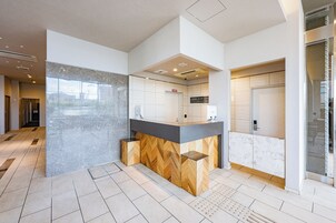 A modern hotel reception area with a marble counter, a herringbone wood accent, and a glass door leading to the elevator.