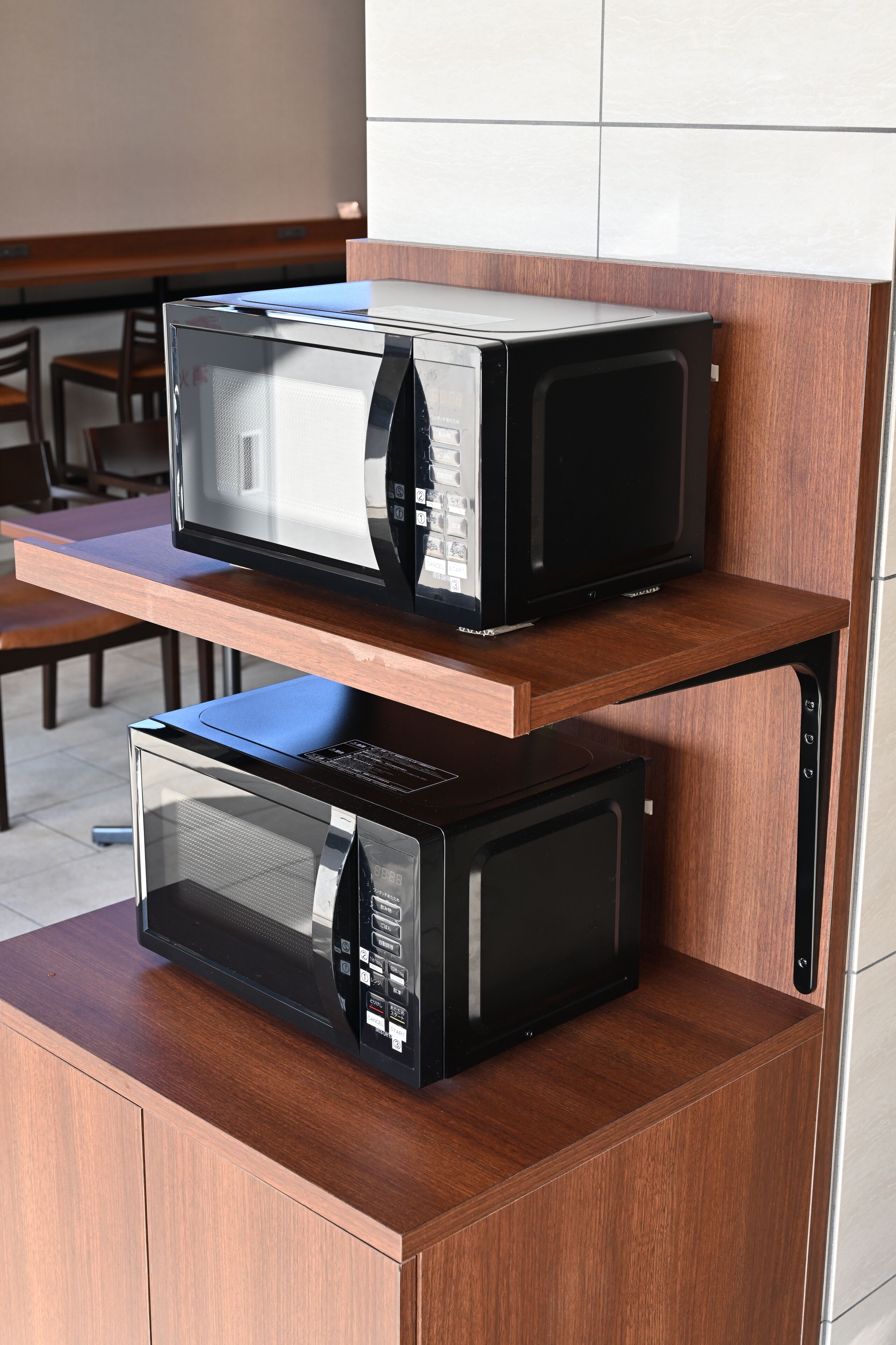 Two black microwaves on a wooden shelf in a kitchen.