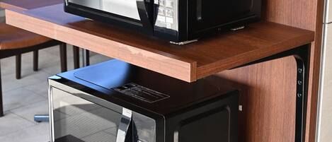 Two black microwaves on a wooden shelf in a kitchen.