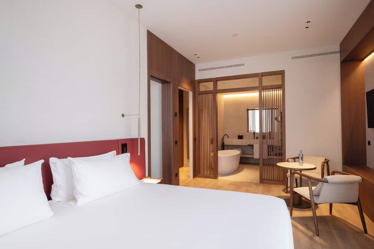 A modern hotel room with a large bed, a small table with chairs, a separate bathroom with a bathtub, and a wooden partition.