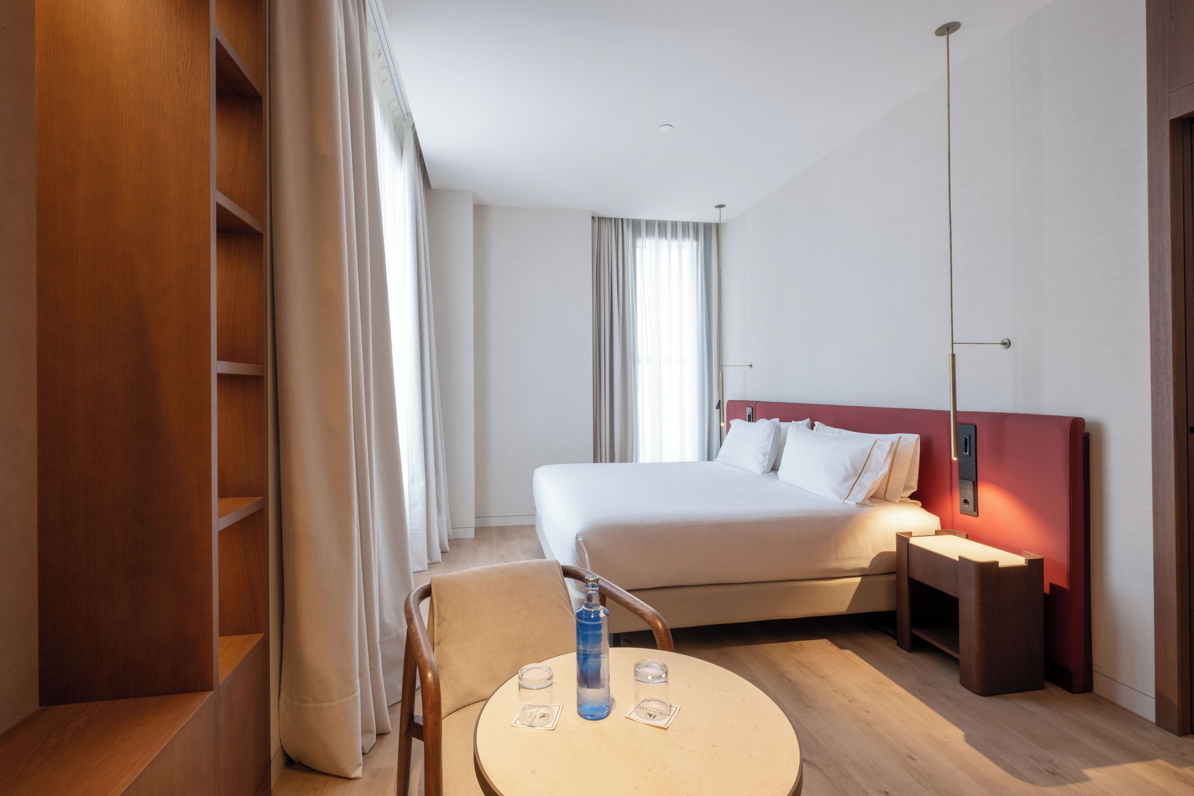A hotel room with a bed, a round table with two glasses, a bottle, and a chair.