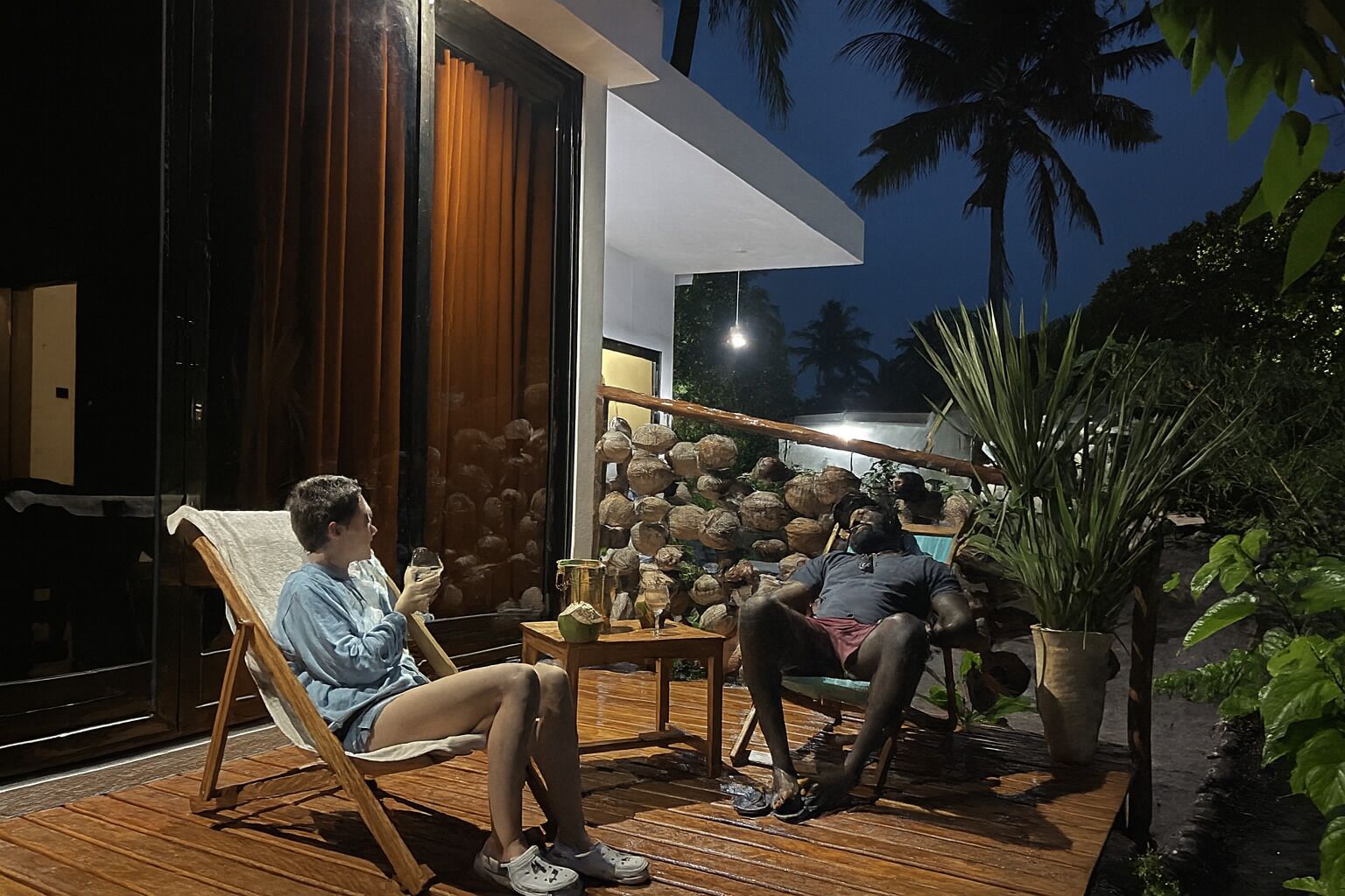 A wooden deck with two people seated, one holding a glass. A large potted plant is visible to the right.