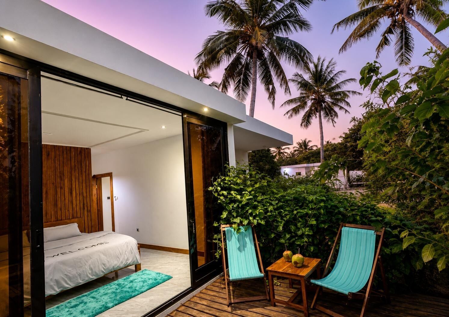 Un dormitorio moderno con cama y puerta corrediza de vidrio que da a un balcón con vistas a palmeras.