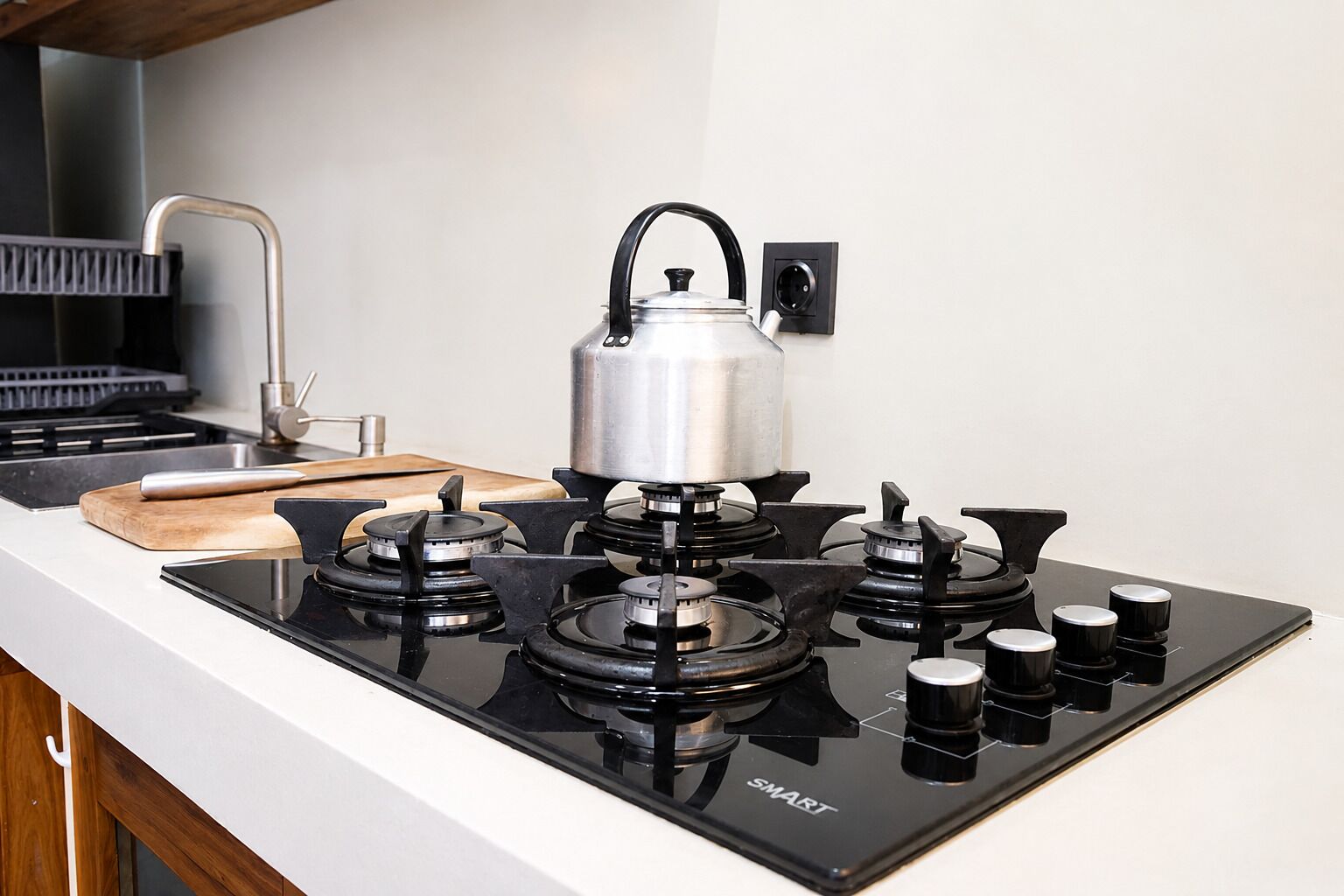 A kitchen with a gas stove, a kettle, and a wooden cutting board.
