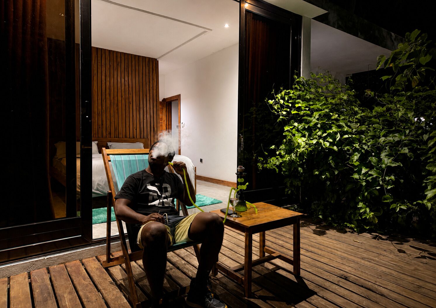 Une personne est assise sur une chaise, sur une terrasse en bois, avec une petite table et un pot de fleurs à proximité.