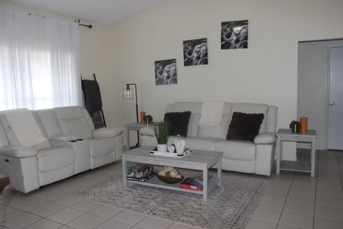 A modern living room with a white sofa set, a coffee table, and a small side table. There are three framed pictures on the wall.