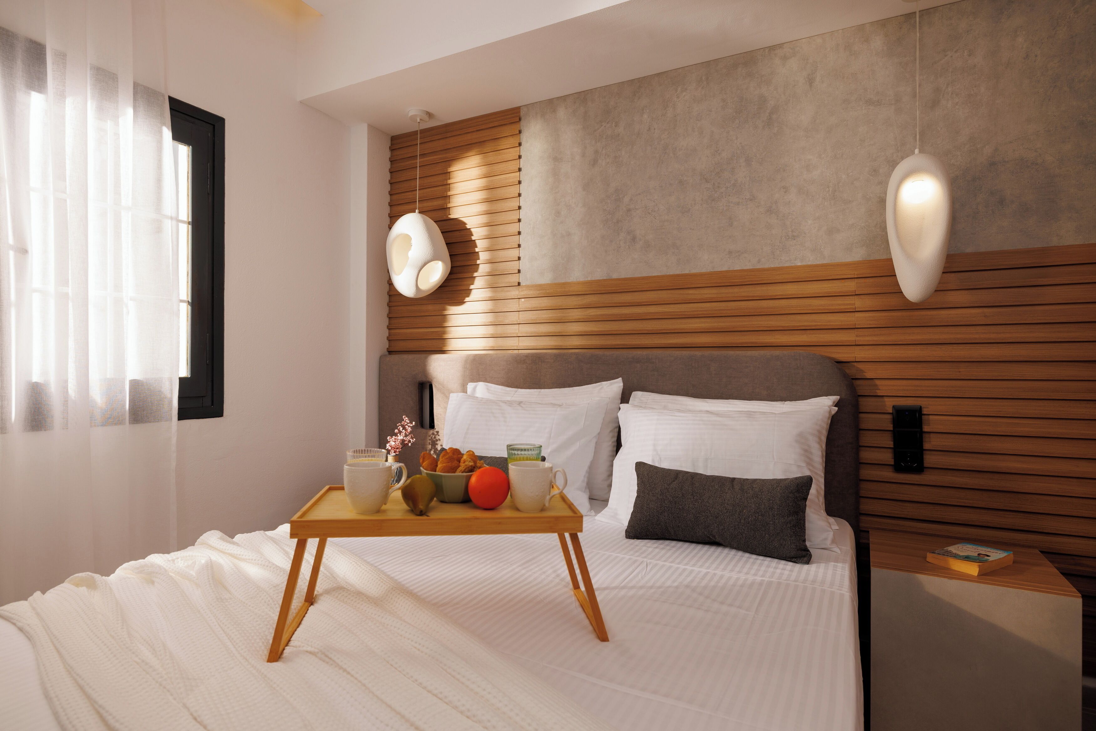 A modern bedroom with a wooden headboard, a bedside table with a book, and a tray with breakfast items.