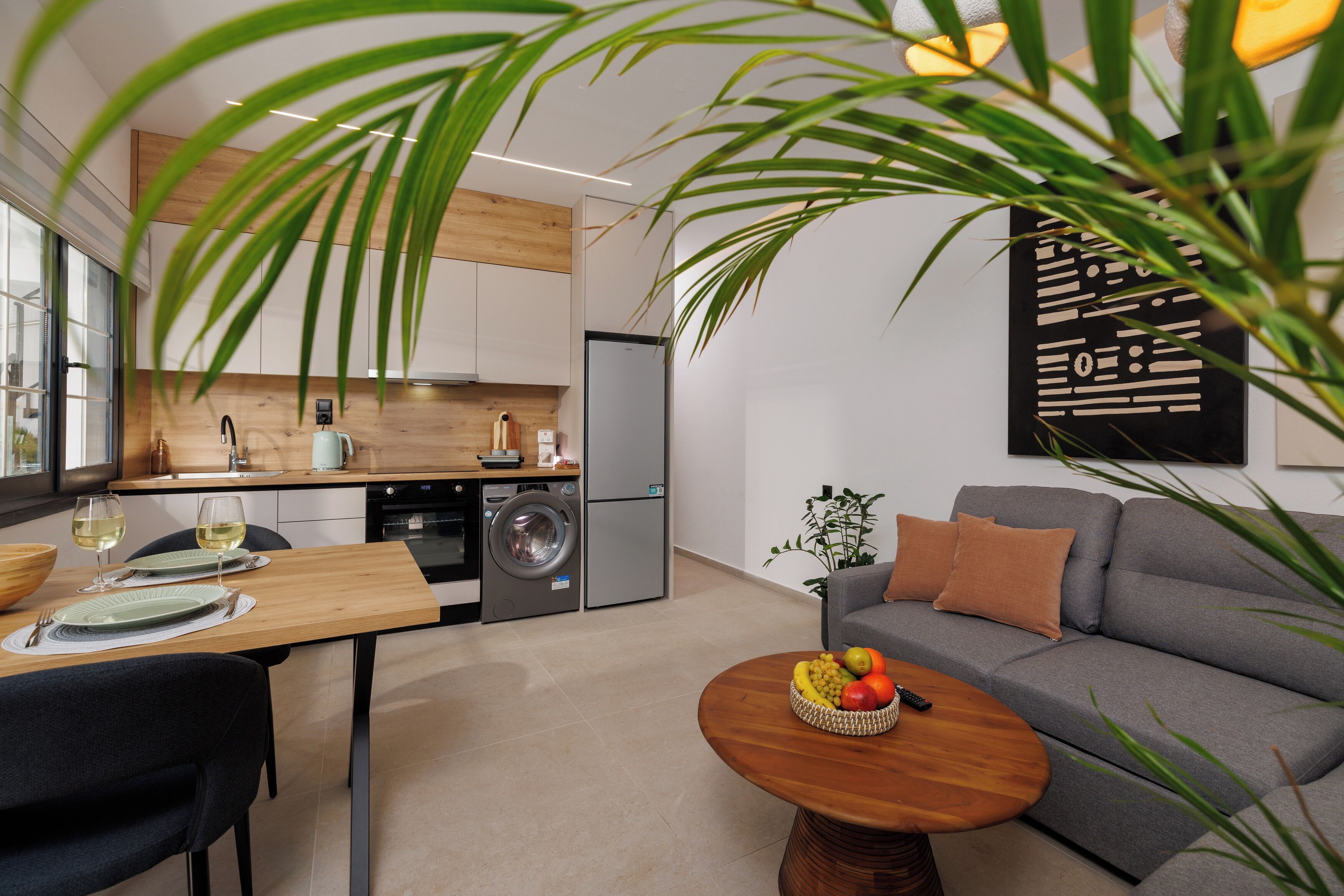 A modern kitchen with a dining area, a grey sofa, and a coffee table with fruit.