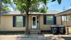 A single-story house with a beige exterior, a front door, and two windows with shutters. There are two trash bins outside. - Home Away From Home! (Amarillo)