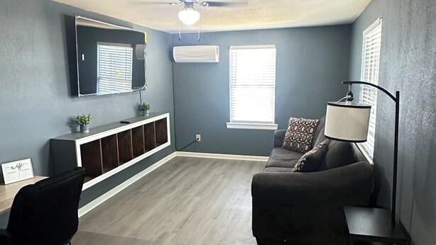 A spacious living room with a dark blue wall, a ceiling fan, a sofa, a coffee table, a TV mounted on the wall, and a window with blinds. - Home Away From Home! (Amarillo)