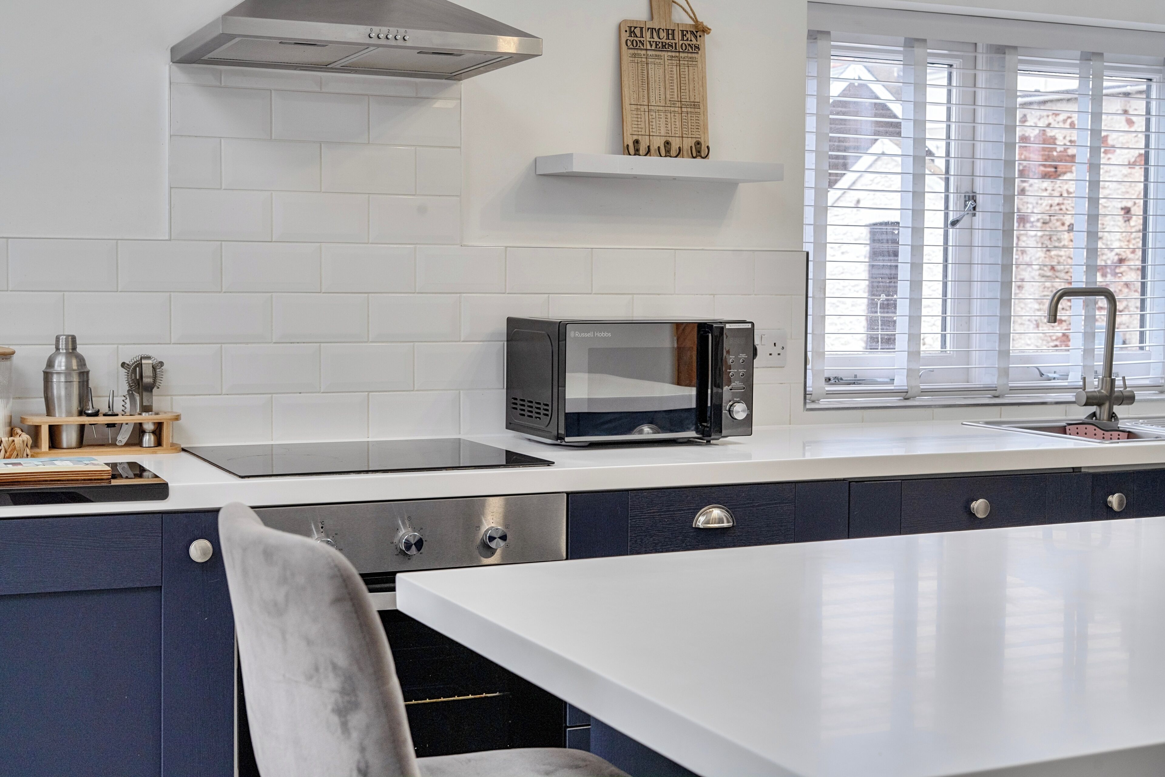 A modern kitchen with white tiles, a built-in microwave, and a window with blinds.