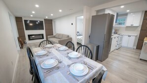 A modern dining area with a table set for four, a sofa, and a kitchen with white cabinets and a stainless steel refrigerator. - 6BR Waterfront Escape | Perfect for Multi-Family Groups (Harcourt)
