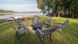 A lakeside outdoor seating area with Adirondack chairs and a round table, surrounded by grass and trees. - 6BR Waterfront Escape | Perfect for Multi-Family Groups (Harcourt)