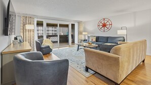 En moderne stue med sofa, lænestole, sofabord og fjernsyn. Der er et stort vindue med gardiner, en vægmonteret kompasdekoration og en skydedør af glas, der fører til et udendørs område.